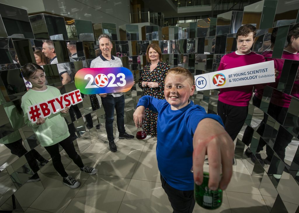 A boy holding a selfie stick with people standing behind with signs for BTYSTE and 2023
